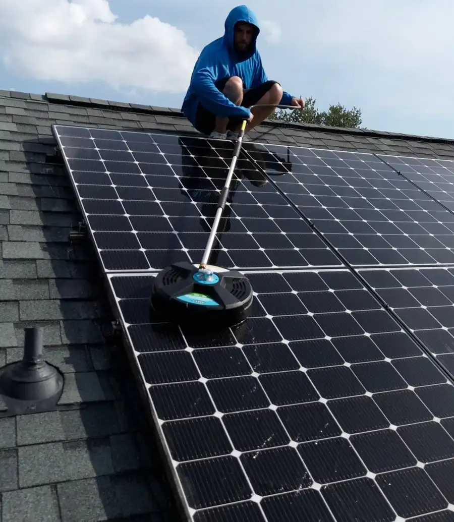 Crew member on a shingle roof cleaning solar panels with a rotating soft-brush head on an extension pole