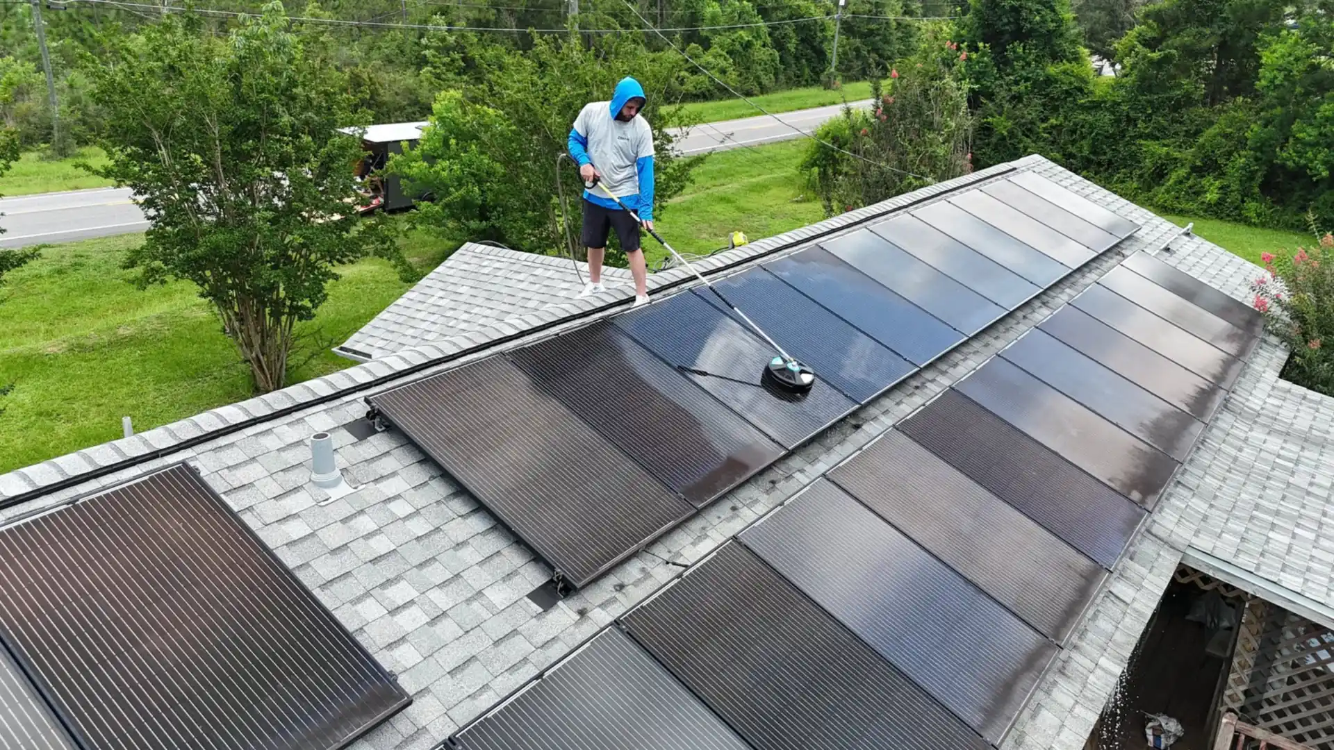 Aerial view of a crew member washing a long row of rooftop solar panels with a soft brush on a water-fed pole