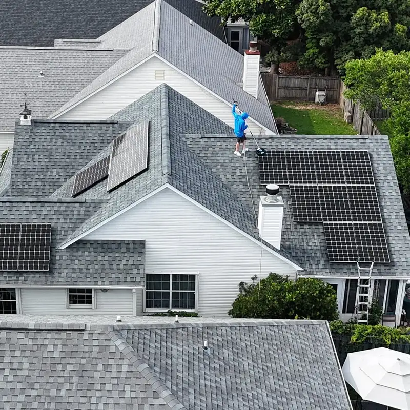 Drone view of a crew member brushing a rooftop solar array on a single-story home, with a ladder against the back wall