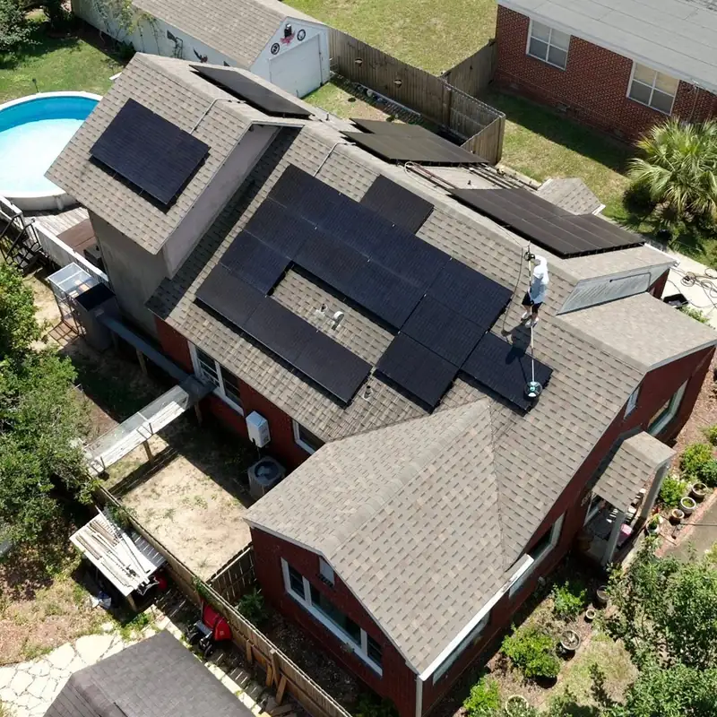 Overhead drone view of a brick home with solar panels across its roof and a crew member cleaning them from the ridge